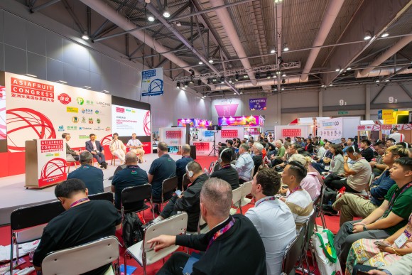 An exhibition hall full with many people sitting in chairs and watching people who speak on the stage. 