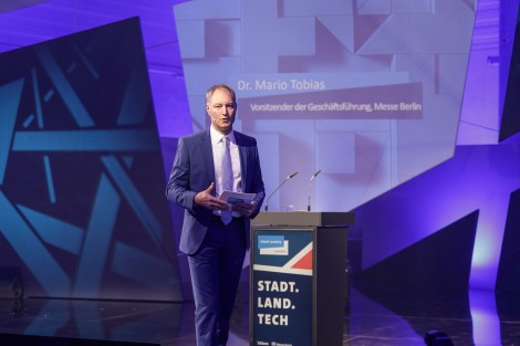 Mario Tobias moderiert auf einer in blau gehaltenen Bühne der Smart Country Convention. Er hat Moderationskarten in den Händen. 